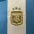 A nova camisa titular feminina da Argentina 2024 destaca o branco com três listras verticais azul celeste. Detalhes azuis estão presentes nas mangas, gola. Logos Adidas e AFA em dourado e o patch de campeã mundial no peito.