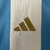 A nova camisa titular feminina da Argentina 2024 destaca o branco com três listras verticais azul celeste. Detalhes azuis estão presentes nas mangas, gola. Logos Adidas e AFA em dourado e o patch de campeã mundial no peito.