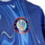 Camisa titular do Chelsea 24/25 é azul royal com padrão de ondas claras, simbolizando chamas. Gola redonda azul, punhos brancos e laranja. Escudo e logo Nike em azul celeste com contorno laranja.