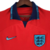 A camisa reserva da Inglaterra para a Copa do Mundo 2022 é vermelha, com gola polo azul e gráfico de 3 Leões. Logo Nike e escudo em azul.