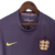 Manto reserva da seleção inglesa com roxo predominante e faixas laterais multicoloridas. Swoosh Nike dourado à direita, Three Lions roxo e dourado à esquerda. Inspirada na camisa reserva da Inglaterra para a Euro 2024.