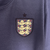 Manto reserva da seleção inglesa com roxo predominante e faixas laterais multicoloridas. Swoosh Nike dourado à direita, Three Lions roxo e dourado à esquerda. Inspirada na camisa reserva da Inglaterra para a Euro 2024.