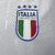 Manto reserva da Itália destaca as cores da bandeira: template branco, listras verdes e vermelhas. Gola azul e branca, frase 'L’Itália Chiamò' na nuca. Logo Adidas azul à direita, escudo FIGC branco à esquerda.