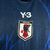 Camisa titular do Japão azul marinho com gráfico de chamas azul royal. Detalhes pretos nos arcos e triangulares. Três listras brancas nas mangas. Escudo da federação japonesa de futebol no peito, logo Y-3 em branco acima.