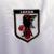 Camisa reserva do Japão 2022: branca com origamis azuis e vermelhos nas mangas. Gola V em azul marinho, escudo da Federação à esquerda, logo Adidas em azul marinho à direita. 