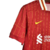 Camisa titular do Liverpool 2024-2025 vermelha com linhas amarelas formando 'YNWA'. Gola branca com friso vermelho e amarelo. Mangas listradas, punhos vermelhos. Escudo Liver Bird e swoosh Nike amarelos.