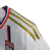 Camisa titular Lyon: branco predominante, faixa vertical vermelha e azul royal no lado esquerdo. Gola V tricolor, três listras douradas nas mangas. Logo Adidas dourado no peito direito, escudo Lyon à esquerda. Manto representa herança e cultura.