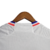 Camisa titular Lyon: branco predominante, faixa vertical vermelha e azul royal no lado esquerdo. Gola V tricolor, três listras douradas nas mangas. Logo Adidas dourado no peito direito, escudo Lyon à esquerda. Manto representa herança e cultura.