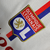 Camisa titular Lyon: branco predominante, faixa vertical vermelha e azul royal no lado esquerdo. Gola V tricolor, três listras douradas nas mangas. Logo Adidas dourado no peito direito, escudo Lyon à esquerda. Manto representa herança e cultura.