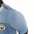 Camisa titular Manchester City 23/24 versão jogador: Azul celeste com faixas verticais em tom sobre tom, inspiradas nas torres do Etihad Stadium. Gola V branca com frisos azul celeste. Escudo do clube e logos PUMA em branco.