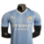 Camisa titular Manchester City 23/24 versão jogador: Azul celeste com faixas verticais em tom sobre tom, inspiradas nas torres do Etihad Stadium. Gola V branca com frisos azul celeste. Escudo do clube e logos PUMA em branco.