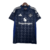 A nova camisa reserva do Manchester United 24/25 traz base azul escuro com padrão de "M" formando estrelas. Detalhes em azul e três listras em prata nos ombros. Escudo e logo Adidas em prata.