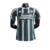 Camisa reserva Manchester United 23/24 versão jogador: Verde escuro com faixas verticais brancas e linhas vermelhas. Gola branca, três listras nos ombros em branco. Escudo Man United verde e branco, logo Adidas branco.