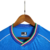 Camisa titular Napoli 23/24: Azul celeste, escudo "N" em jacquard, gola V cruzada com cores da Itália, detalhes brancos, escudo e logo EA7. MSC no patrocínio máster, patch campeão italiano.