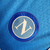 Camisa titular Napoli 23/24: Azul celeste, escudo "N" em jacquard, gola V cruzada com cores da Itália, detalhes brancos, escudo e logo EA7. MSC no patrocínio máster, patch campeão italiano.