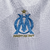 Camisa titular Olympique de Marseille 2023: branco com tons de cinza inspirados na rocha calcária, gola e punhos azul celeste com linha branca, logo PUMA dourado, escudo Marseille no lado esquerdo.