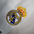 Camisa titular Real Madrid branca com gola em V e frisos em azul marinho, branco e dourado. Escudo no peito e logo Adidas em azul marinho. Frase "¡Hala Madrid!" na nuca.