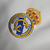 Camisa titular Real Madrid branca com gola em V e frisos em azul marinho, branco e dourado. Escudo no peito e logo Adidas em azul marinho. Frase "¡Hala Madrid!" na nuca.