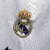 Camisa titular manga longa Real Madrid branca com gola em V e frisos em azul marinho, branco e dourado. Escudo no peito e logo Adidas em azul marinho. Frase "¡Hala Madrid!" na nuca.