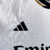 Camisa titular manga longa Real Madrid branca com gola em V e frisos em azul marinho, branco e dourado. Escudo no peito e logo Adidas em azul marinho. Frase "¡Hala Madrid!" na nuca.