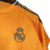 A camisa reserva do Real Madrid tem cor laranja predominante com linhas poligonais estilizadas. Detalhes em preto na gola, listras dos ombros, escudo e logos da Adidas e Emirates.