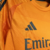 A camisa reserva do Real Madrid tem cor laranja predominante com linhas poligonais estilizadas. Detalhes em preto na gola, listras dos ombros, escudo e logos da Adidas e Emirates.