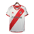 Camisa branca do River Plate com faixa diagonal vermelha. Detalhes em vermelho na gola V, três listras, punhos, laterais e barra. Escudo Millonarios na faixa à esquerda, logo Adidas vermelho à direita. 'Grandeza' na nuca .