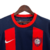 Camisa titular do San Lorenzo 2024-2025 azul marinho com duas faixas verticais vermelhas. Gola redonda e punhos vermelhos, ombros e mangas azul marinho. Escudo San Lorenzo com círculo central maior no peito esquerdo, logo Nike branco no direito.