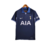 Camisa reserva do Tottenham 2023-2024 em azul marinho com gola pólo lilás claro, inspirada no código postal N17. Logos Nike e escudo iridescentes, detalhes em ondas nas mangas e corpo.