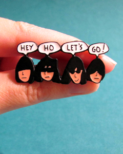 Pin de metal com desenho do rosto dos integrantes da banda Ramones dizendo "hey ho let's go!". Possui dois fechos na parte de trás.