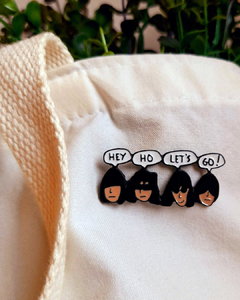 Pin de metal com desenho do rosto dos integrantes da banda Ramones dizendo "hey ho let's go!". Possui dois fechos na parte de trás.