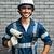 Hombre sonriente con casco y gafas de seguridad sosteniendo una cámara de vigilancia, vestido con overol azul y guantes, frente a una pared de ladrillos.