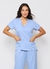 Esta imagem mostra uma modelo vestida de scrub feminino azul claro celeste, que esta à venda. O uniforme consiste em uma blusa de manga curta com decote em V e dois bolsos frontais, e uma calça combinando. O conjunto é ideal para clínica, hospital e home 