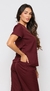 Modelo vestida com scrub feminino bordô Vinho, que esta à venda, ela esta de frente. O scrub é composto por uma blusa de manga curta com decote em V e dois bolsos frontais, e uma calça combinando. 