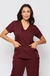 Modelo vestida com scrub feminino Bordô lindo que esta à venda, ela esta de frente. O scrub é composto por uma blusa de manga curta com decote em V e dois bolsos frontais, e uma calça combinando. 