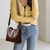 Mulher usando uma bolsa de ombro de couro marrom, com um suéter amarelo e jeans, destacando o estilo e a elegância.