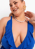 Modelo plus size sorrindo com um vestido azul de decote profundo e detalhes em babados, usando colar de pérolas e brincos em forma de folha.