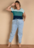 Modelo vestindo blusa solta em malha com decote canoa e recortes contrastantes, combinada com calça jeans clara e sandálias, sorrindo em fundo neutro.