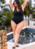 Modelo plus size usando maiô preto em poliflex com decote redondo e drapeado, posando ao lado de uma piscina.