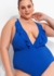 Mulher plus size sorrindo usando um maiô azul com detalhes em babados, posando com um brinco elegante e um colar de pérolas.