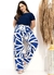 Modelo plus size sorridente usando blusa azul e calça larga estampada em azul e branco, posando ao lado de um vaso decorativo.