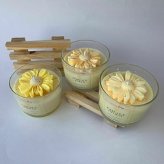 Três velas decorativas em copos de vidro com flores feitas de cera de coco nas cores amarelo e creme, acompanhadas de um suporte de madeira.