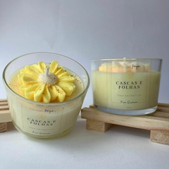 Duas velas aromáticas da coleção "Cascas e Folhas", uma com decoração de flor amarela e a outra com um design mais simples.