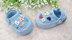 Mini crocs de crochê azul para bebê com letras coloridas decorativas.