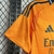 Imagem da camisa reserva do Real Madrid 24/25 em laranja com detalhes metálicos azuis.