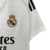 Imagem da camisa titular Real Madrid 24/25, com design branco tradicional, gola em V e detalhes em dourado e azul marinho.