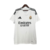 Imagem da camisa titular Real Madrid 24/25, com design branco tradicional, gola em V e detalhes em dourado e azul marinho.