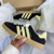 Tênis ADIDAS GAZELLE INDOOR na cor preto com detalhes em amarelo, modelo com três listras laterais e sola translúcida.