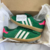 Um par de tênis ADIDAS GAZELLE INDOOR na cor verde, com detalhes em rosa e três listras brancas, dentro de uma caixa com papel de seda.