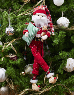 Muñeco de Santa Claus con pijama de puntos rojos y bata, sosteniendo un oso de peluche y almohada  en sus brazos.